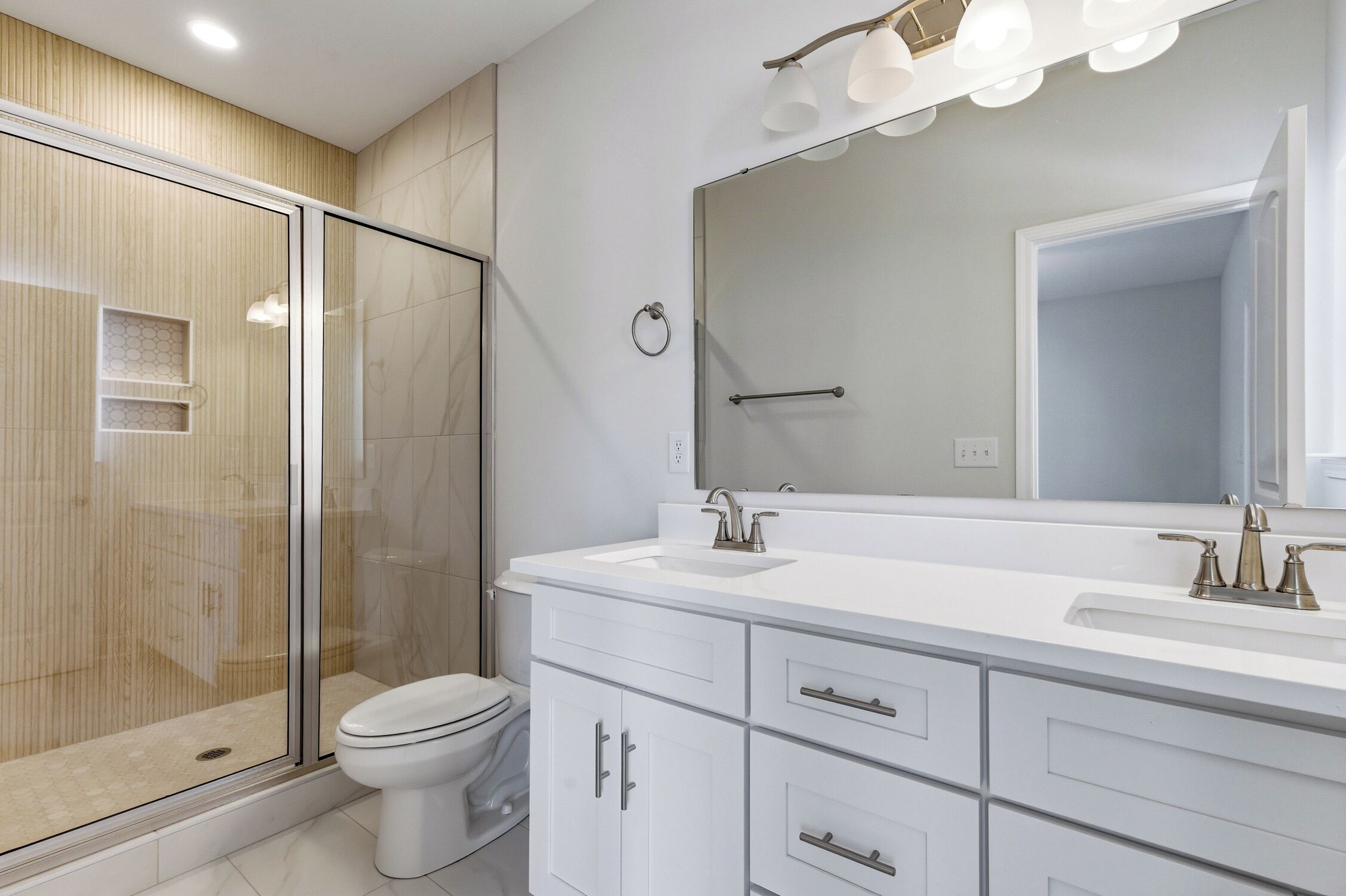 A modern bathroom with a glass shower enclosure, a white toilet, a large mirror with lights, and a white vanity with drawers.