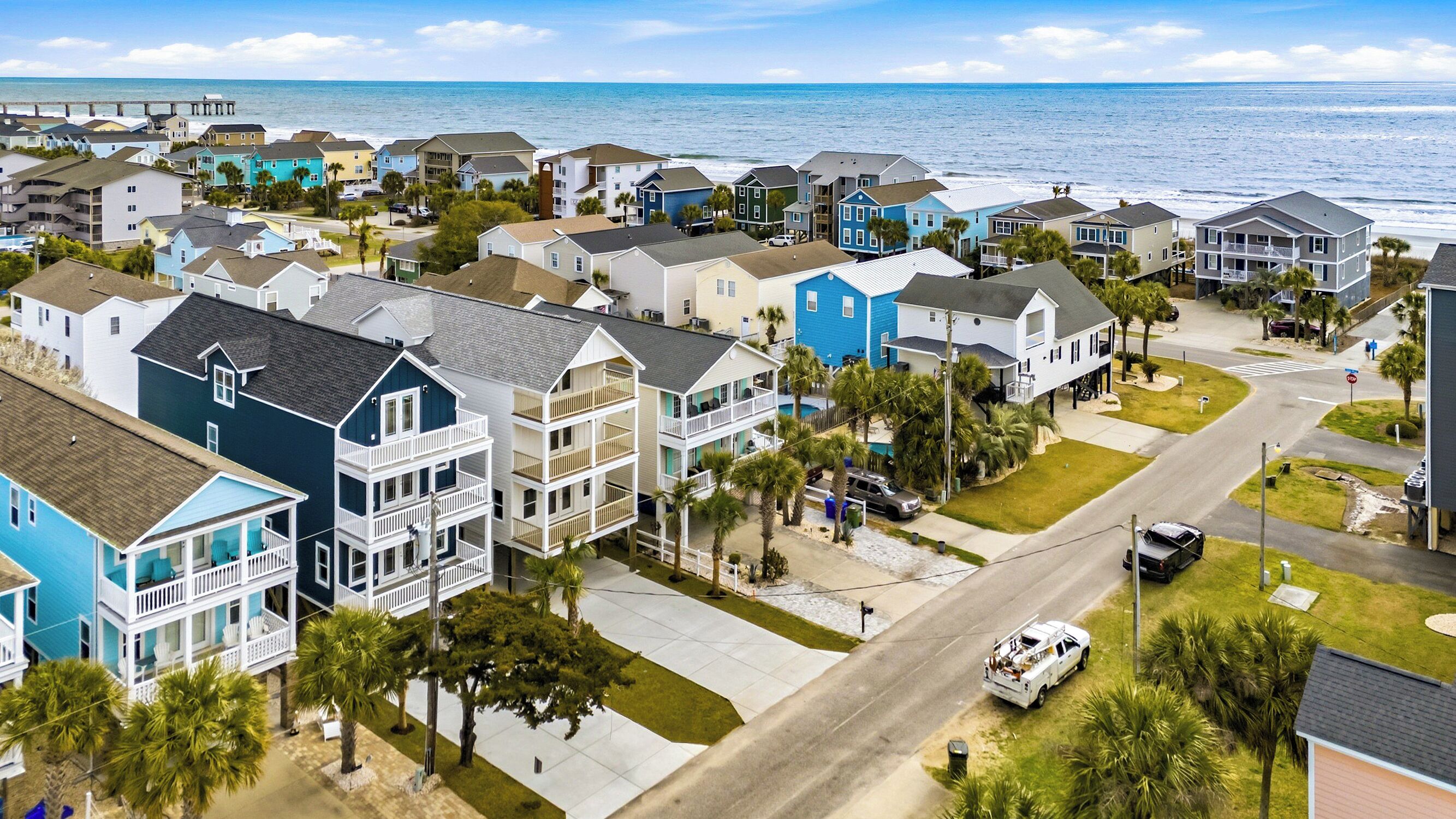 A coastal residential area with multi-story houses, a visible road, and a pier in the background.