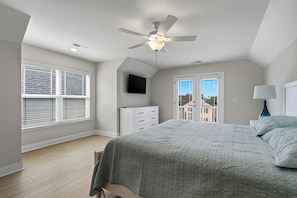 Une chambre à coucher comprenant un lit, une commode, un ventilateur de plafond, un téléviseur et une fenêtre avec des stores.