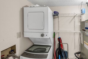 A laundry room with a stacked washer and dryer, a shelf with cleaning supplies, and a wall-mounted electrical box. - Horizon East Cabana 4 - Oceanfront in Garden City Beach (Garden City Beach)