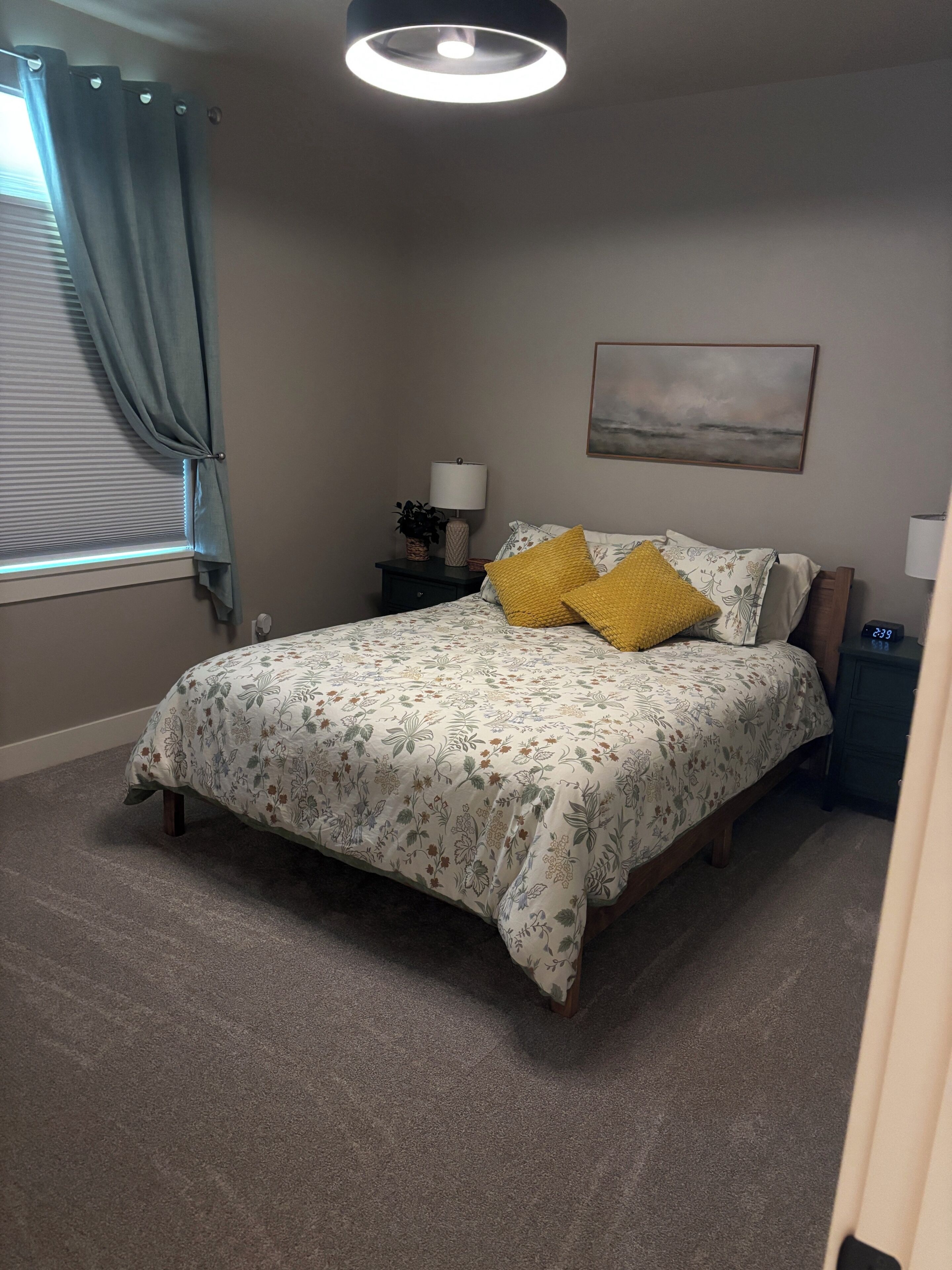 A bedroom with a bed, a nightstand, a lamp, a wall clock, and a window with curtains.