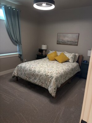 A bedroom with a bed, a nightstand, a lamp, a wall clock, and a window with curtains.