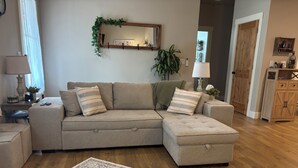 Ein Wohnzimmer mit Ecksofa, einem gemusterten Teppich, einem Spiegel mit Grünpflanzen und einem Holzschrank.