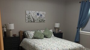 Una camera da letto con un letto, due comodini, una lampada a parete, una finestra con tende e un'opera d'arte floreale al muro.