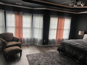 Ein Schlafzimmer mit einem Bett, einem Sessel, einer Lampe und einem Fenster mit durchsichtigen Vorhängen.