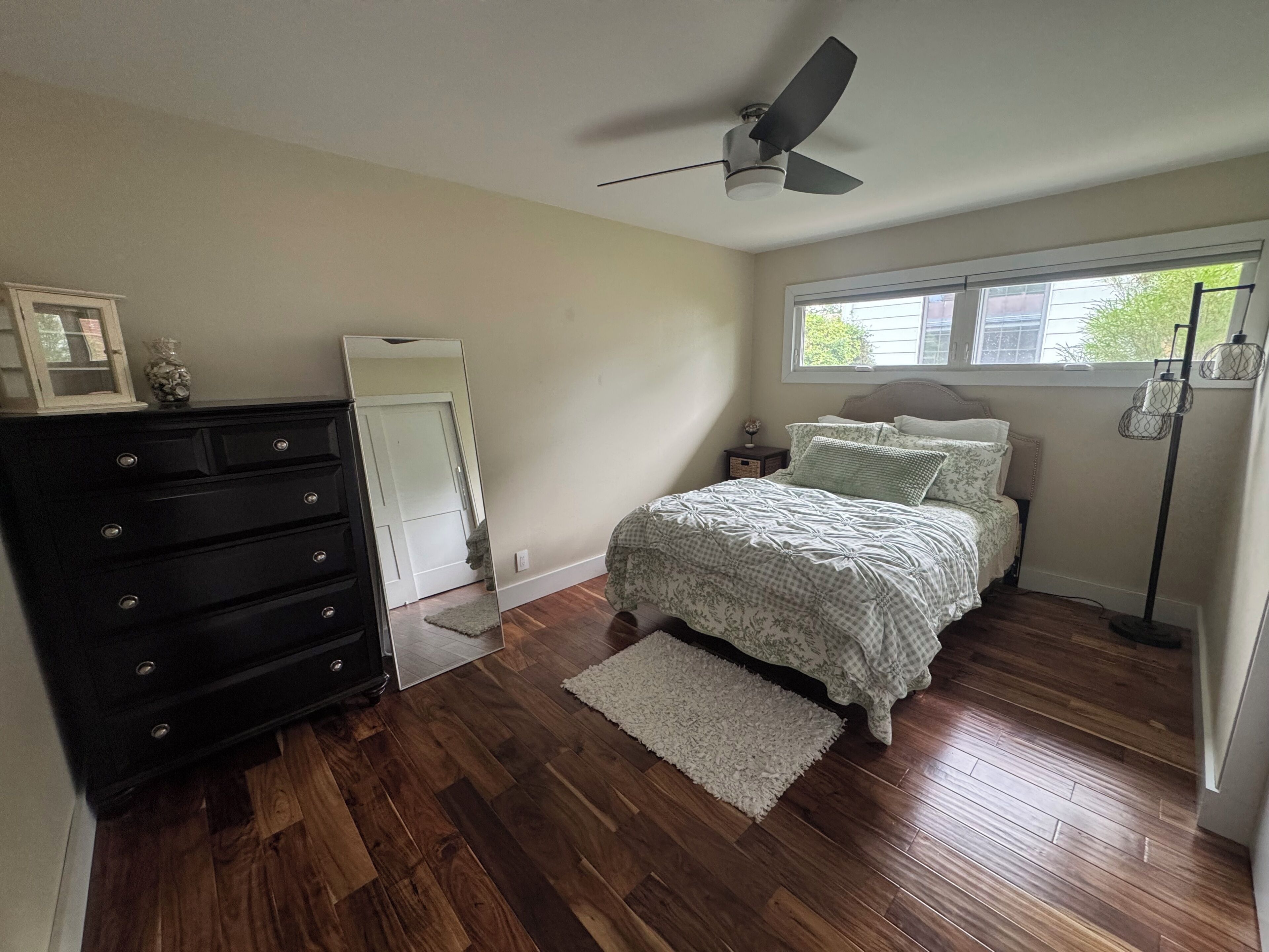 Une chambre à coucher comprenant un lit, une commode, un ventilateur de plafond, une fenêtre et une lampe sur pied.