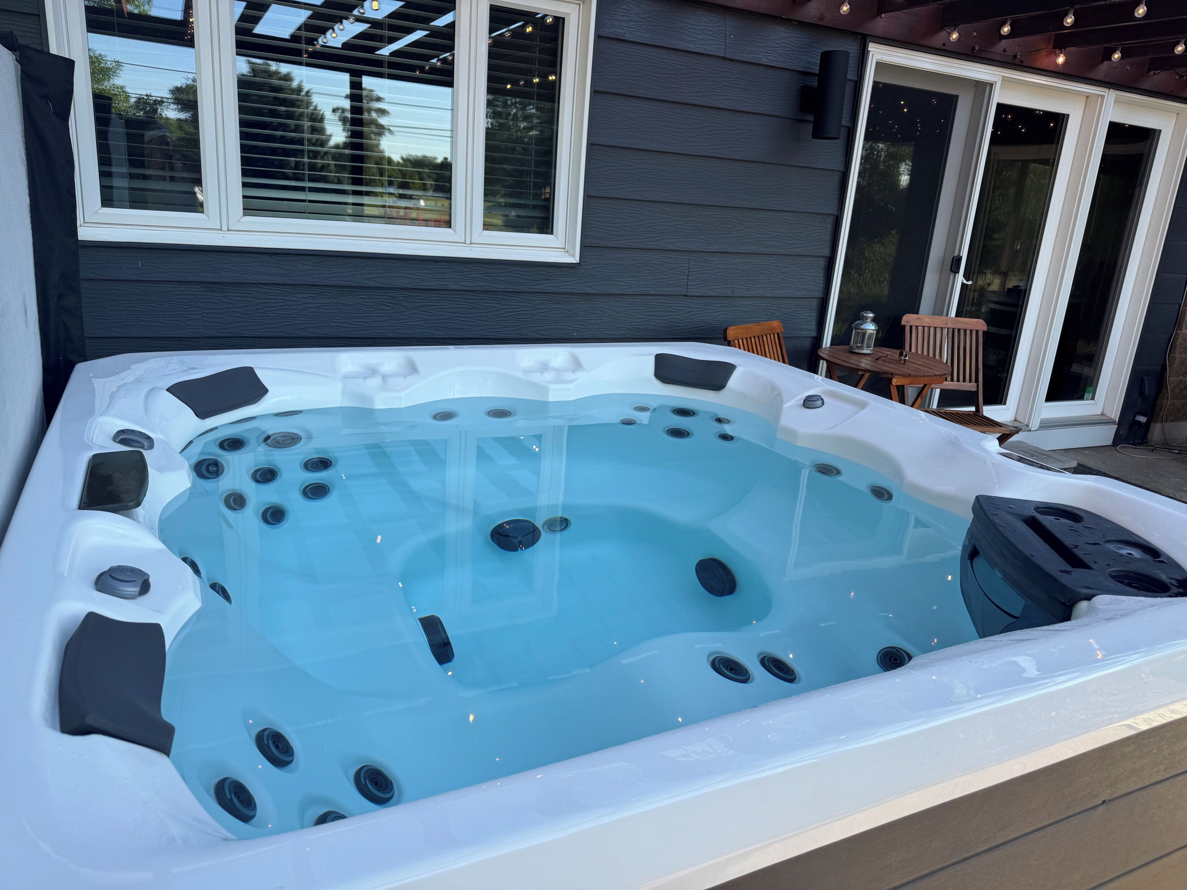 A hot tub with a clear view of the surrounding area, including a house with a pergola and a patio chair.