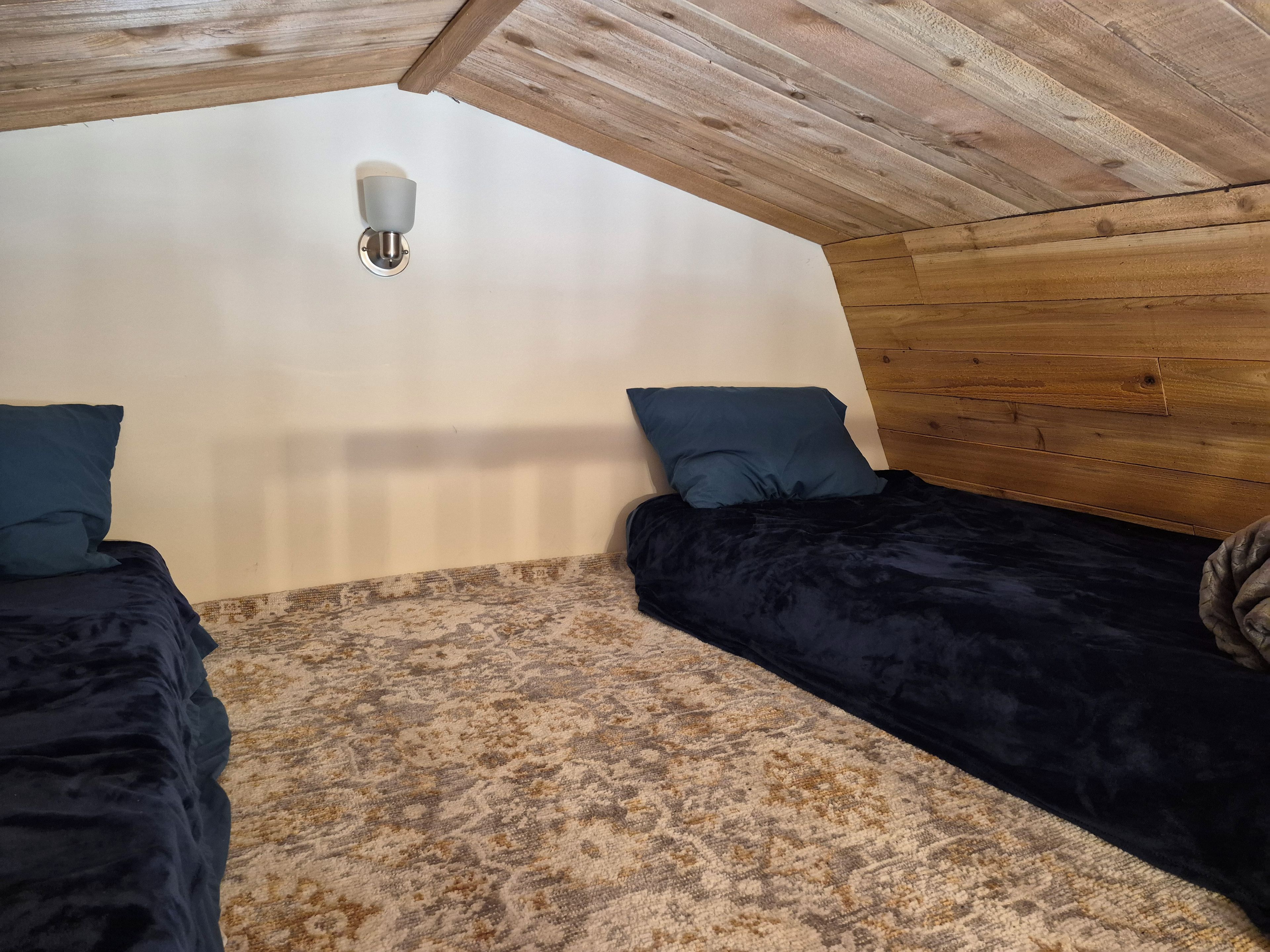 Een slaapkamer met twee bedden, een kamerbreed tapijt, houten balken in het plafond en een wandlamp.