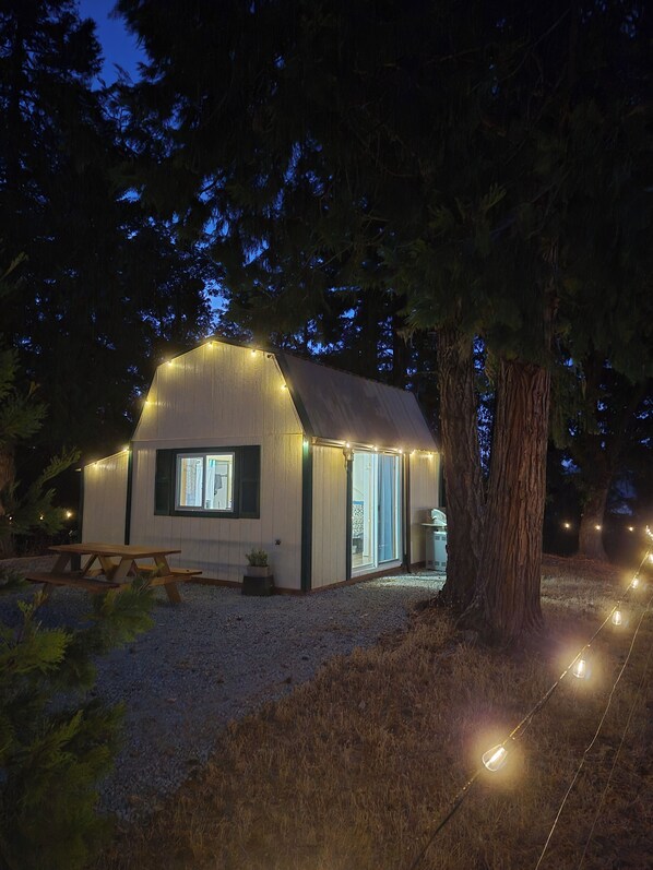 A small cabin with a sloped roof, a wooden picnic table, and a string of lights.