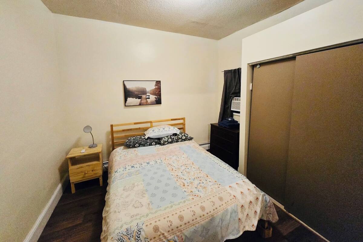 A bedroom with a bed, a nightstand, a picture on the wall, a window with curtains, and a door.