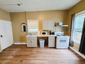 A compact kitchen with a microwave, stove, and sink, a small trash bin, and a window with blinds.
