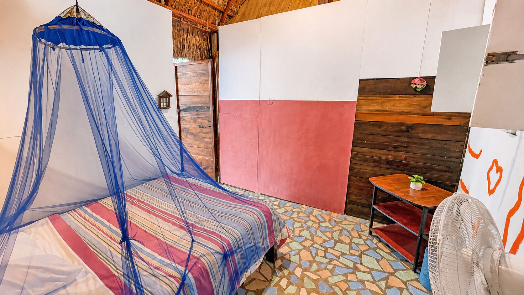 A room with a bed, a fan, a small table with a plant, and a wall with a wooden panel.