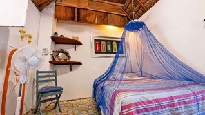 A bedroom with a bed under a blue canopy, a chair, a wall-mounted shelf with decorative items, and a wall-mounted fan. - Hostal & Camping El Árbol de la Vida. (Bacalar)