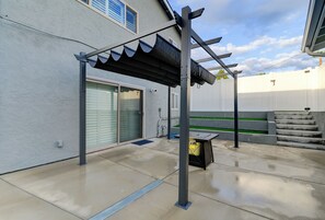 A covered patio area with a pergola structure, a sliding glass door, and a white fence in the background. - Luxury 2BR Guesthouse Near SDSU & Mission Trails (San Diego)