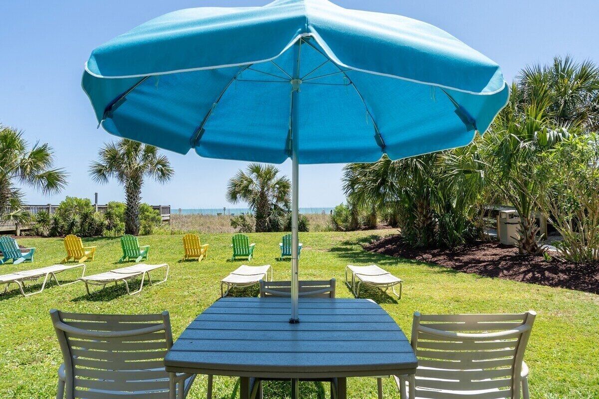 Ein Sitzbereich im Freien mit Tisch und Stühlen unter einem großen Sonnenschirm, umgeben von Palmen und Blick auf das Meer.