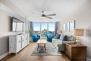 Un salon moderne doté d’un ventilateur de plafond, d’un téléviseur à écran plat, d’une table basse en bois et offrant une vue sur la ville grâce à de grandes fenêtres.