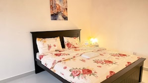 En seng med blomstret sengetøj, et natbord med en lampe og et billede på væggen.