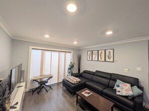 A living room with a black leather sofa, a wooden coffee table, a keyboard, and a large window with blinds. - The Bloom House - 2 Bedroom 2 Bath Family Home (Vancouver)