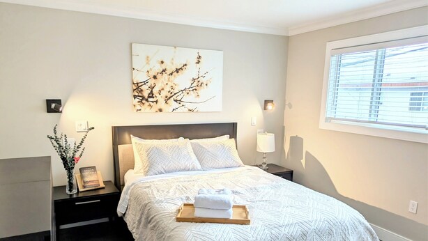 Een slaapkamer met een bed, nachtkastjes, een wandlamp, een raam met jaloezieën en een kunstwerk aan de muur.
