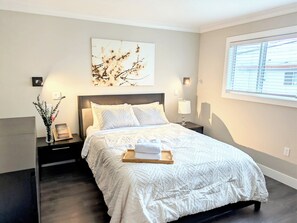 A bedroom with a bed, bedside tables, a nightstand, a wall-mounted lamp, a window with blinds, and a wall art piece. - The Bloom House - 2 Bedroom 2 Bath Family Home (Vancouver)
