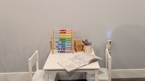 A room with a white table, two white chairs, a wooden toy abacus, a cup with pencils, and a framed abstract artwork on the wall.