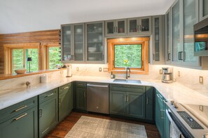 A modern kitchen with dark green cabinets, a built-in dishwasher, and a window with a view of greenery. - Mountain Chic Cabin | Hot Tub, Firepit & Seclusion in Blue River (Blue River)