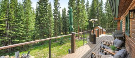 Une terrasse en bois avec des meubles d’extérieur, un foyer et une vue sur une forêt.