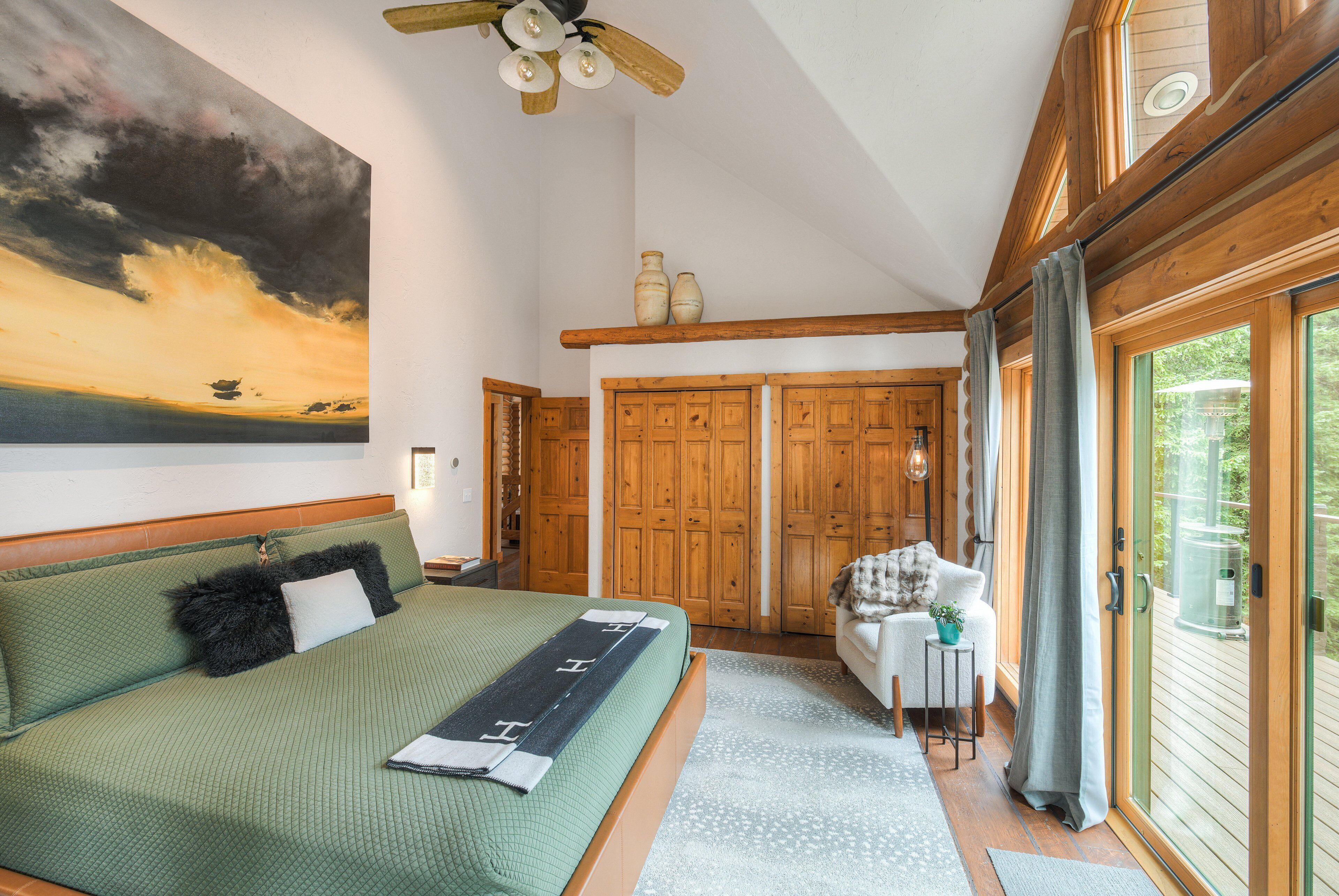Een slaapkamer met een bed, een schilderij, houten deuren en uitzicht door een raam.