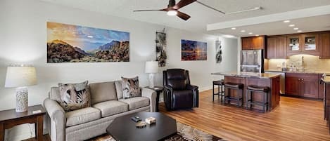 A modern living room with a sofa, coffee table, and a dining area with stools. There is a ceiling fan and artwork on the walls.