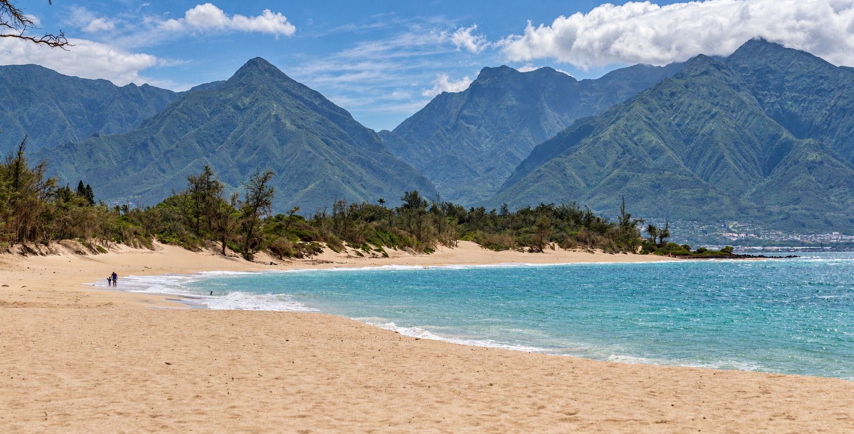 A beach with a mountain backdrop, clear skies, and a few people walking.