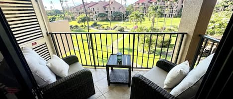 A balcony with wicker furniture, a small table, and a view of a well-maintained lawn and buildings in the background.