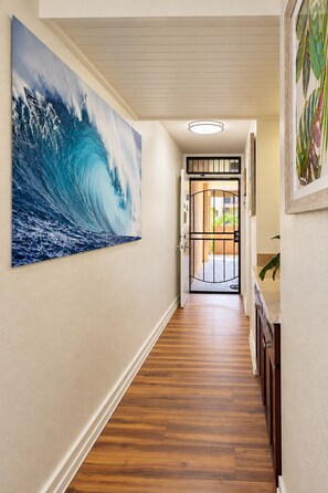 A hallway with a wooden floor, a large wave painting on the left wall, and a door at the end. - South Kihei Luxury Condo • Gorgeous Sunset Views.  Steps to Kamaole Beach III (Kihei)