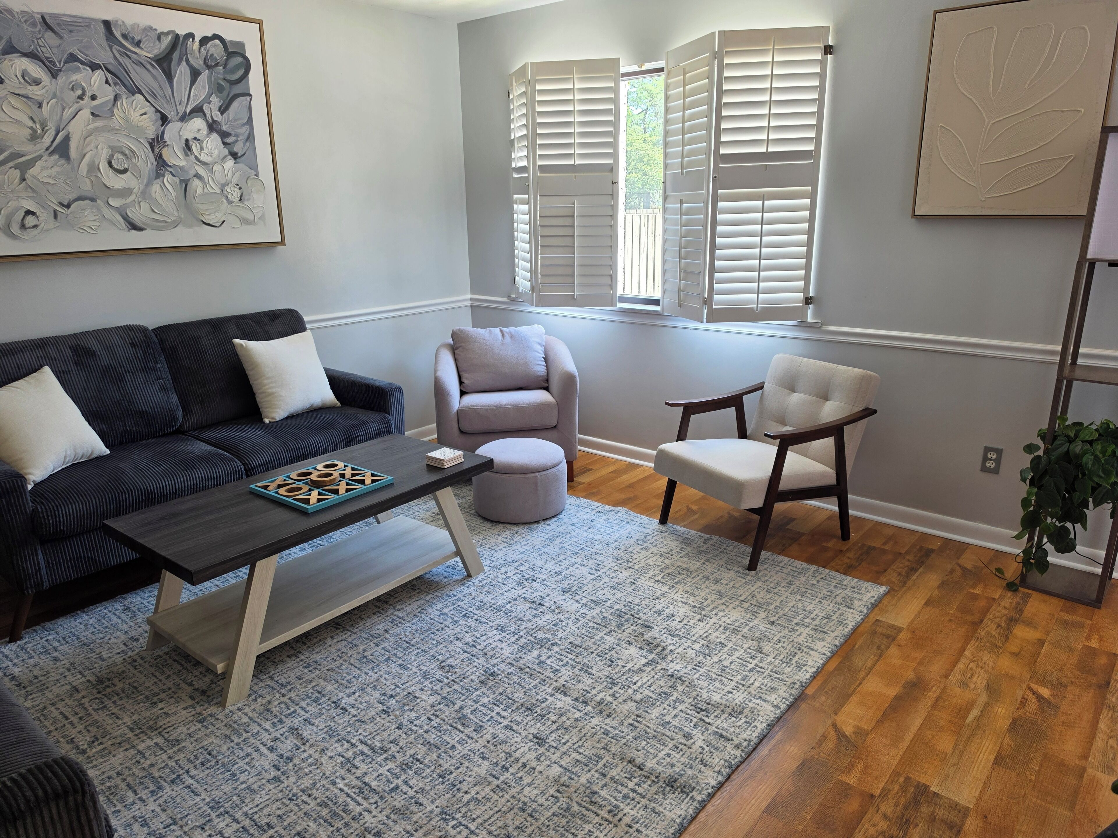 A living room with a dark sofa, a light-colored armchair, a small table with a board game, and a window with shutters.