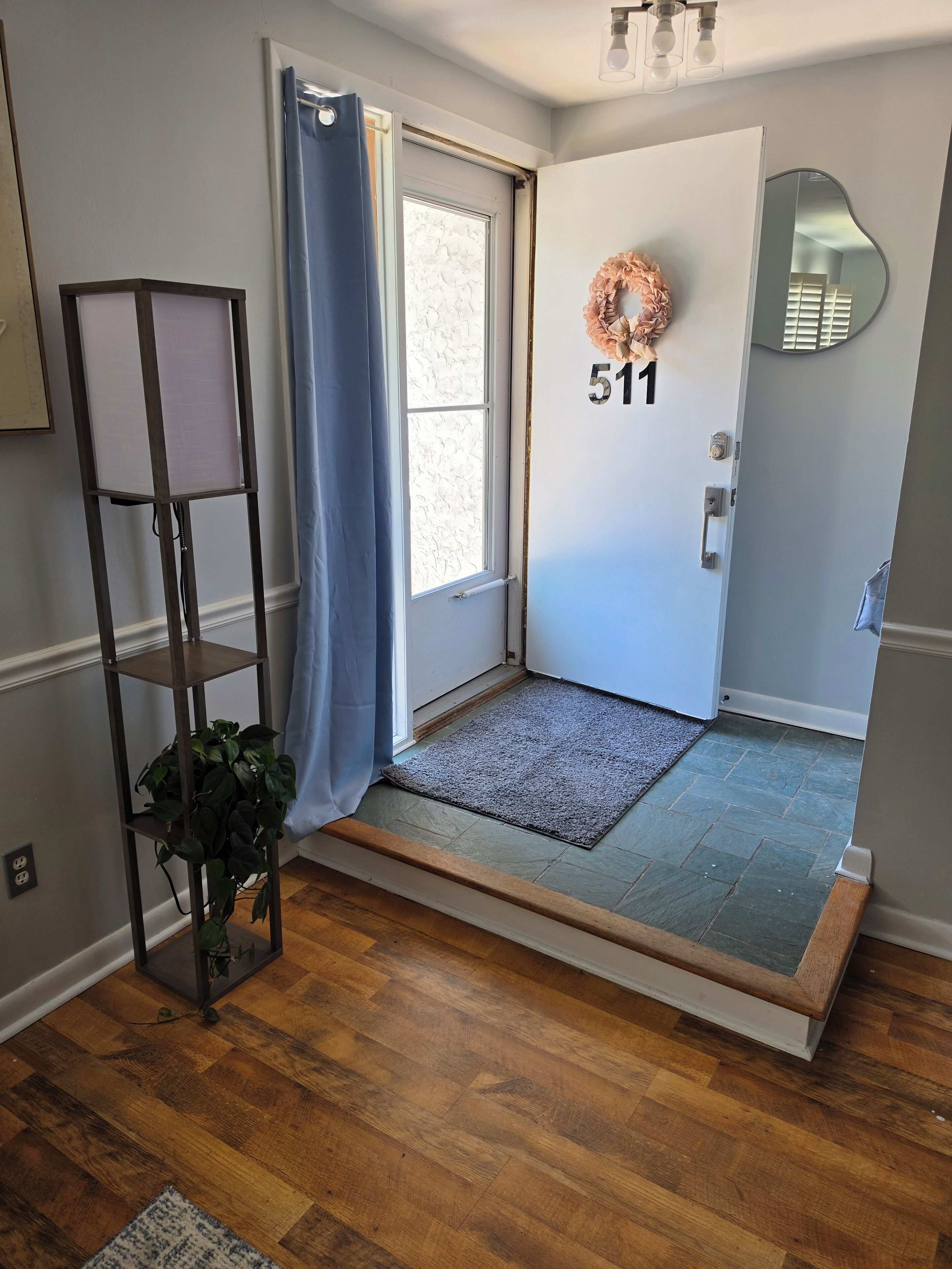 A room with a wooden floor, a door with a number plaque, a blue curtain, a plant on a metal stand, and a mirror on the wall.