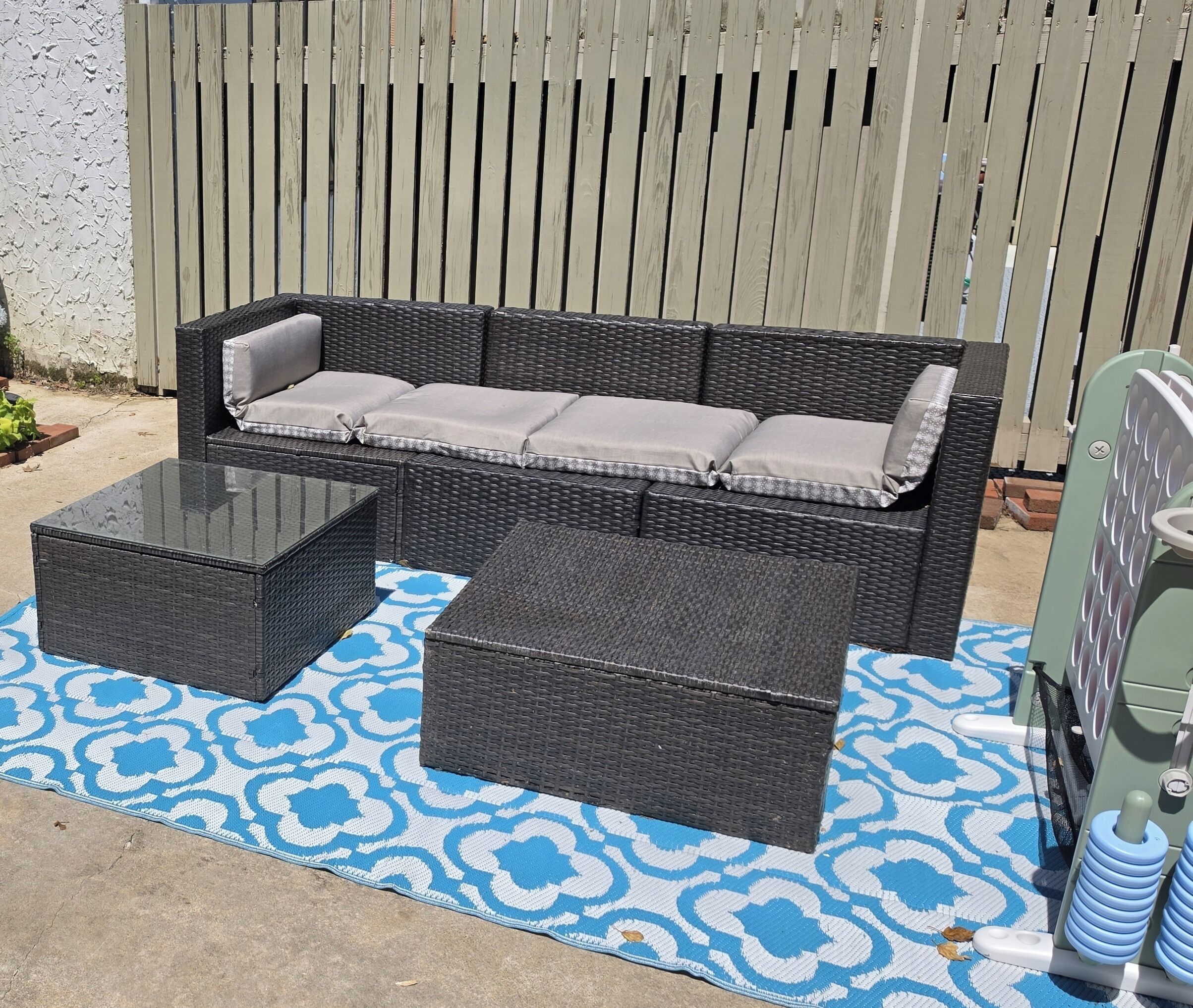 A wicker sofa with cushions, a matching ottoman, and a glass-top coffee table, all set on a patterned outdoor rug.