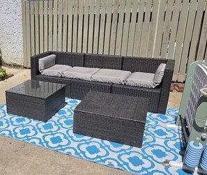 Un canapé en osier agrémenté de coussins, un pouf assorti et une table basse avec un plateau en verre, le tout posé sur un tapis d’extérieur à motifs.