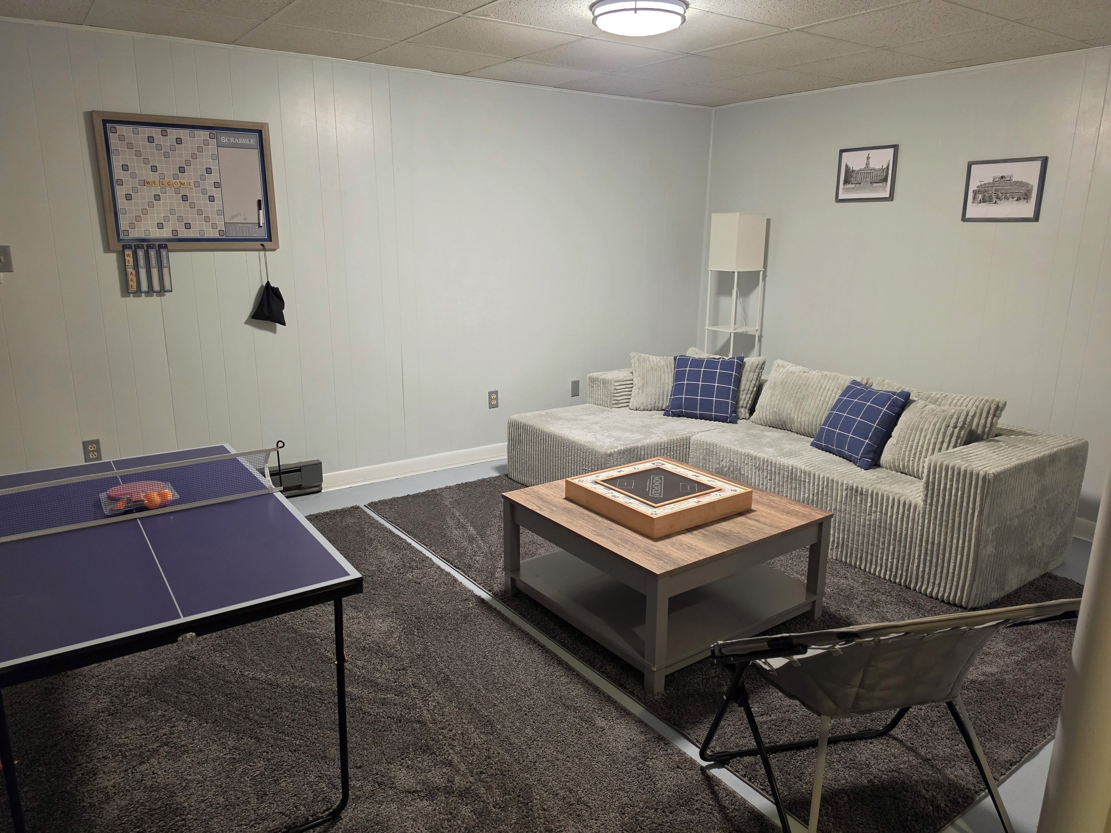 Una habitación con mesa de ping pong, un sofá con cojines, una mesita y cuadros enmarcados en la pared.