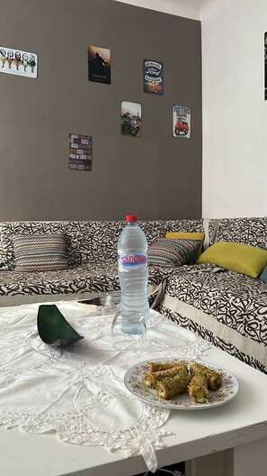 A living room with a patterned sofa, a table with a bottle of water, a plate of food, and wall decorations. - Comfortable Apartment in the Heart of Tunis – Explore the City Easily (Tunis)