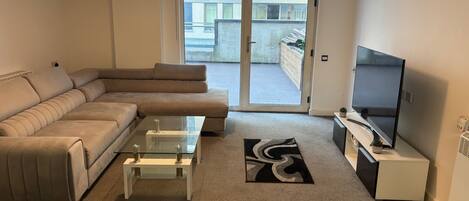 A modern living room with a sofa, a glass coffee table, a flat-screen TV, and a sliding glass door leading to a balcony.