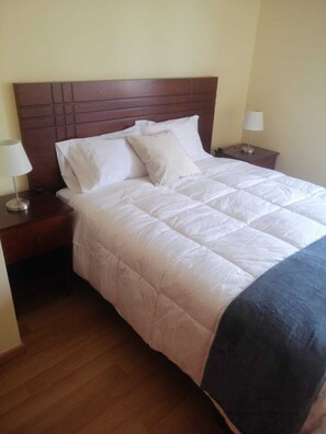 Een net opgemaakt bed met een houten hoofdbord, witte beddengoed en twee nachtkastjes met lampen.