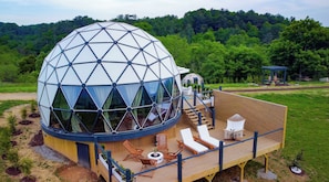 Une structure en forme de dôme géodésique dotée d’une terrasse, de sièges extérieurs et offrant une vue sur une zone boisée.
