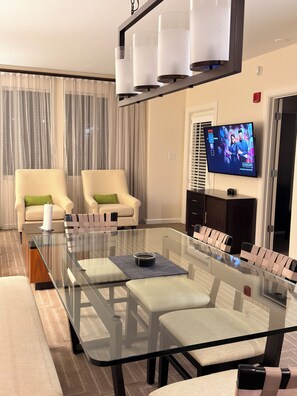 A modern living room with a glass-top dining table, two sofas with green cushions, a flat-screen TV mounted on the wall, and a ceiling light fixture. - King Villa Resort with Balcony near Orlando’s I-Drive (Orlando)