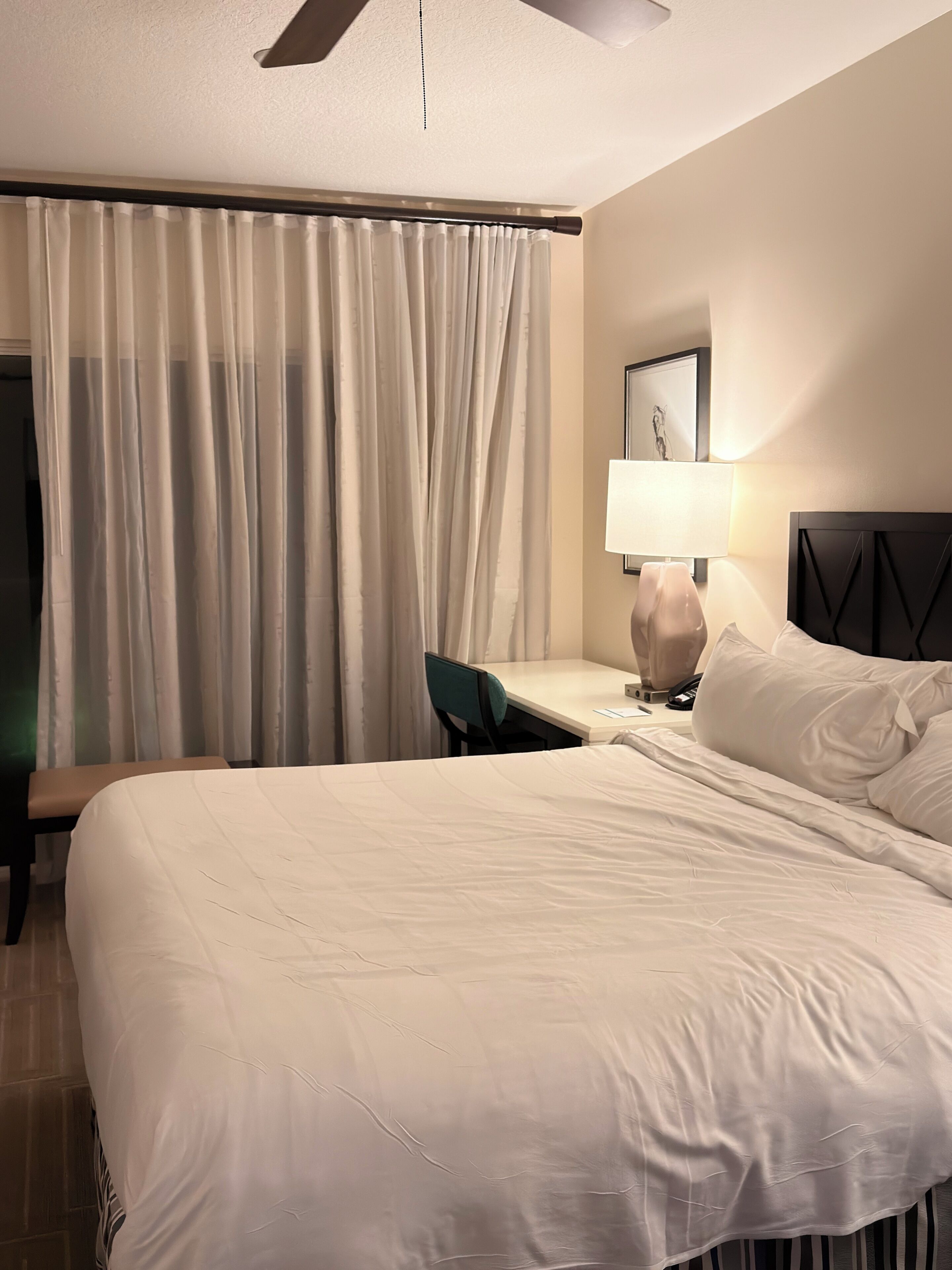 A hotel room with a bed, a desk, a chair, a lamp, and a window with curtains.