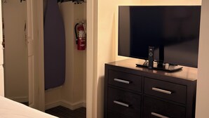 Een hotelkamer met een bed, een televisie op een dressoir, een kast met kleding en een brandblusser.