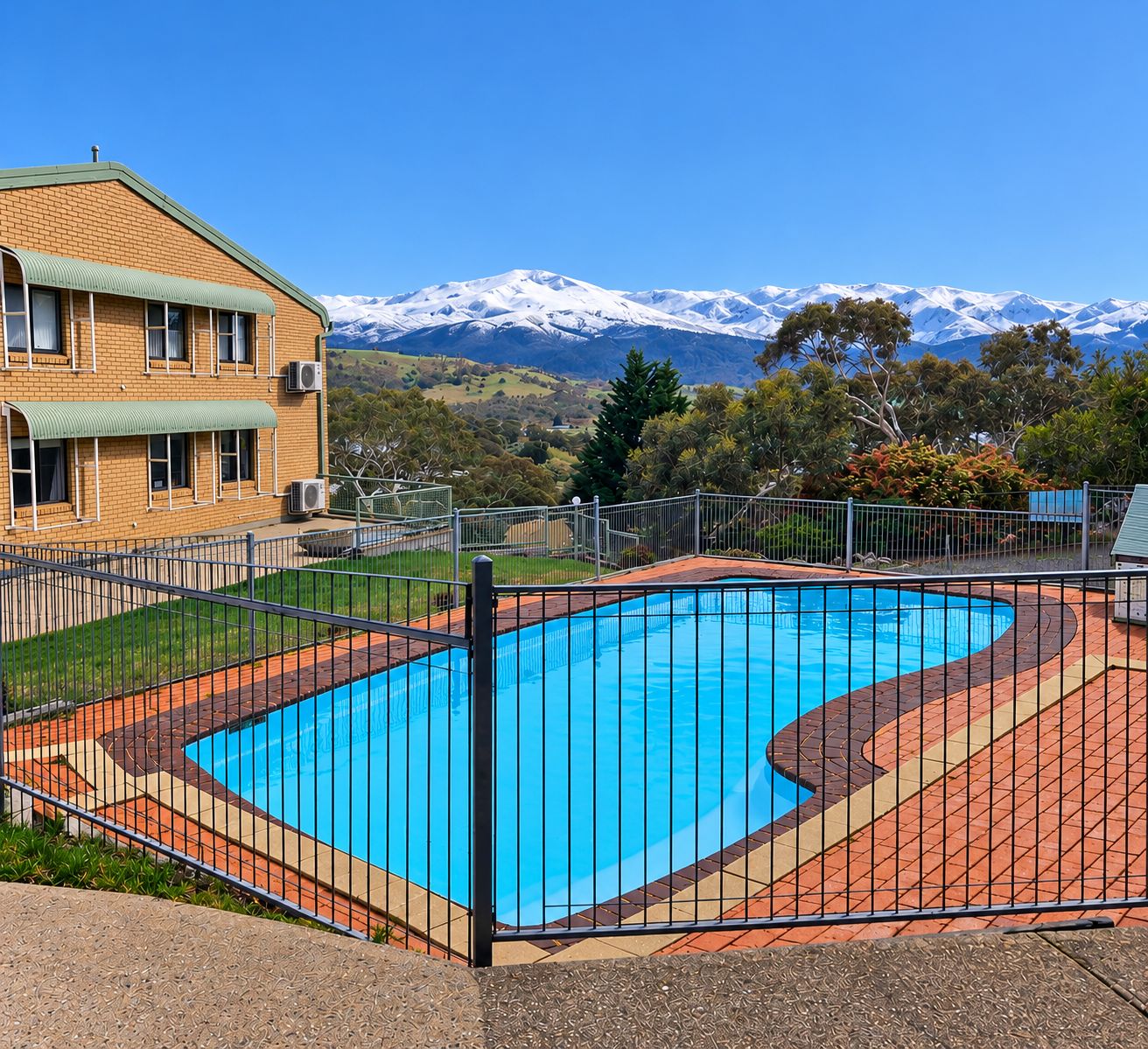 A swimming pool with a curved design, surrounded by a metal fence, and a building with a green roof in the foreground.