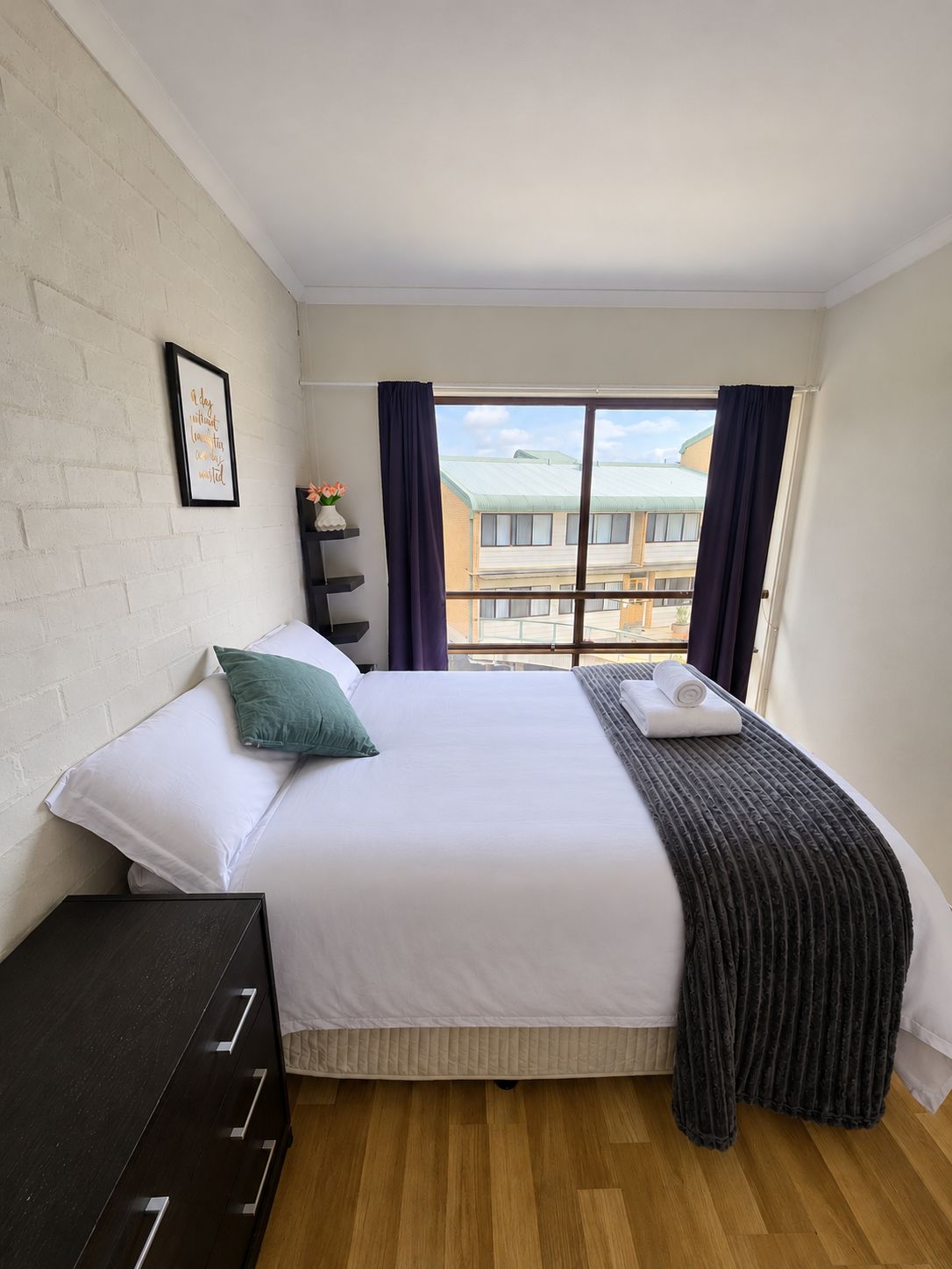 A bedroom with a bed, a dresser, a window with curtains, and a view of buildings outside.