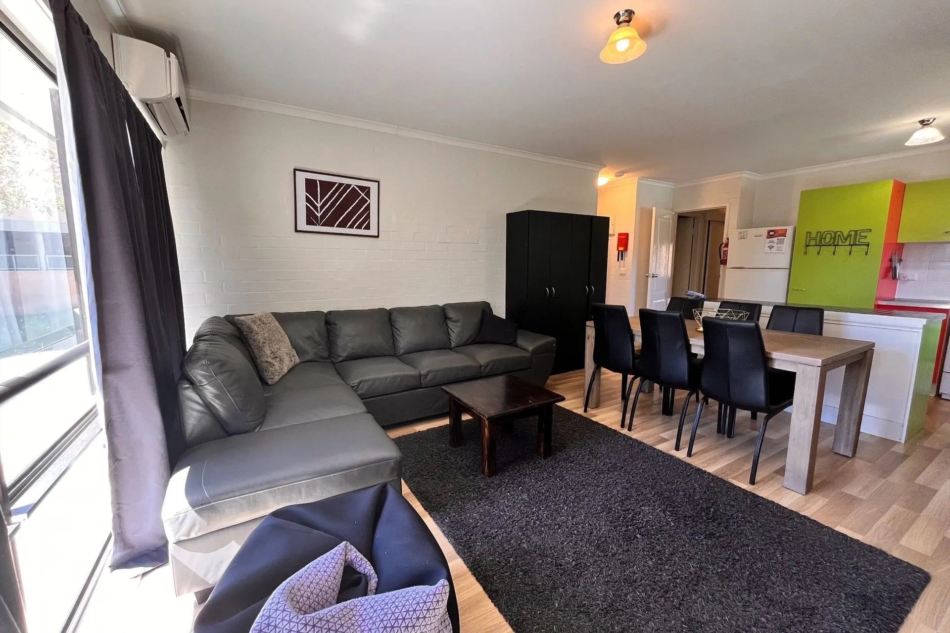 Ein modernes Wohnzimmer mit einem Ecksofa, einem Essbereich mit Stühlen, einer Küche mit Kühlschrank und einem Fenster mit Vorhängen.