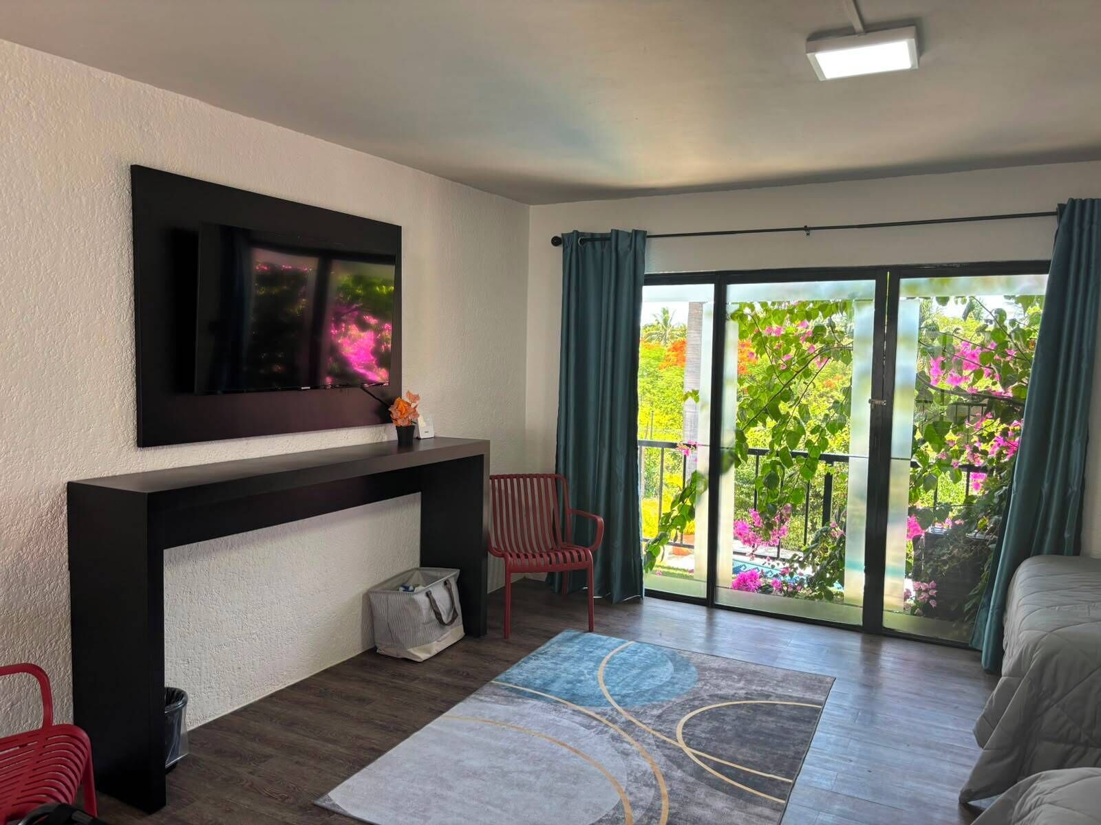 A modern hotel room with a large sliding glass door to a balcony, a flat-screen TV mounted on the wall, a red chair, and a bed with a white duvet.