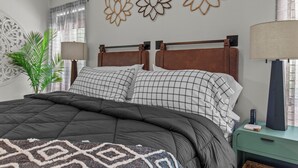 A neatly made bed with a dark quilt, two pillows with a checkered pattern, and a decorative wall with three floral wall art pieces.