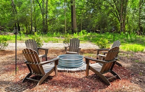 A circular fire pit with four Adirondack chairs around it, set on a bed of mulch in a forested area.
